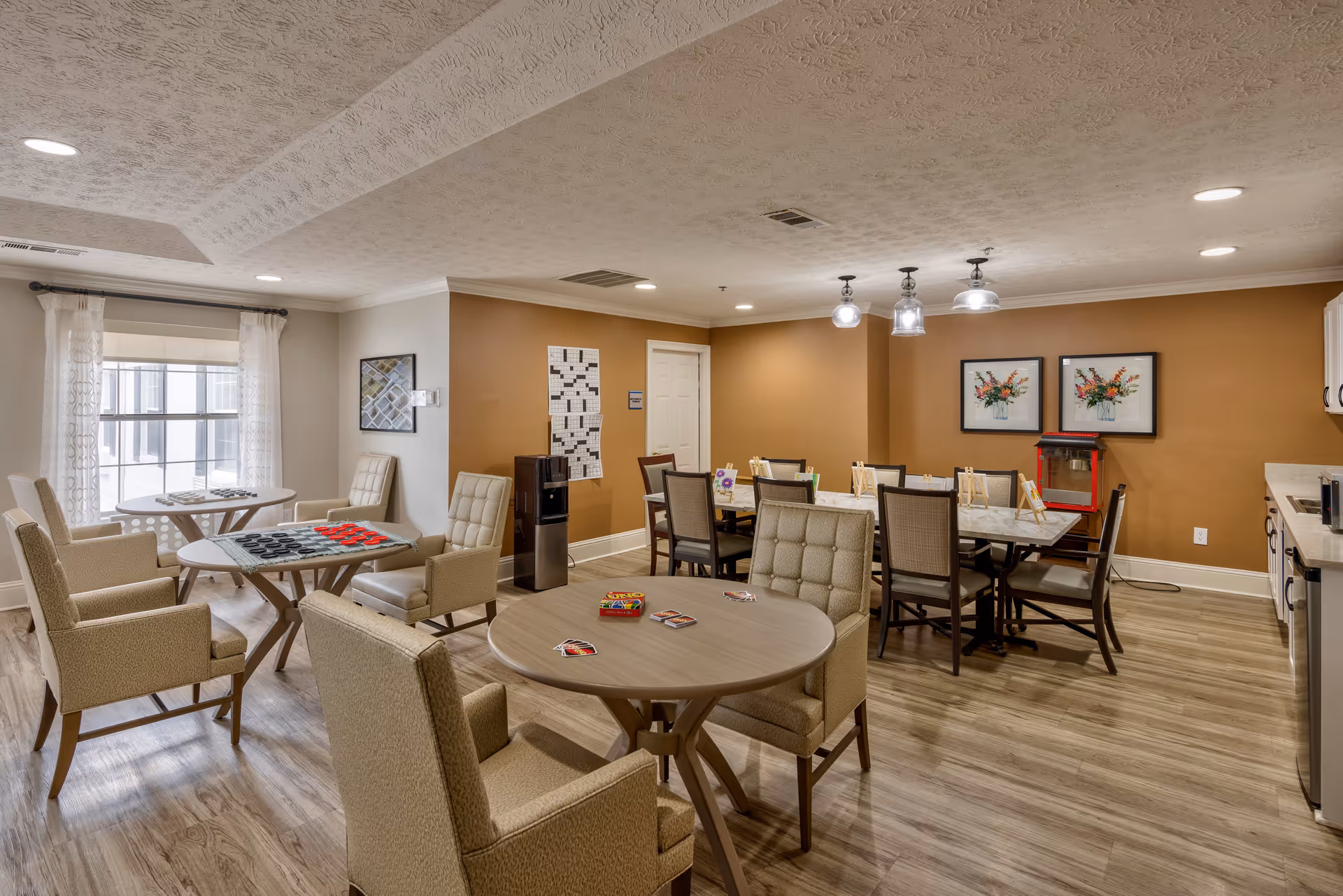 A well-lit common area in a senior living facility with multiple round tables and cushioned chairs arranged for social activities. One table has a checkers game set up, another has cards, and there are additional tables and chairs in the background near a popcorn machine and framed floral artwork on the wall. The room has wood flooring, beige walls, and a window with sheer curtains letting in natural light.