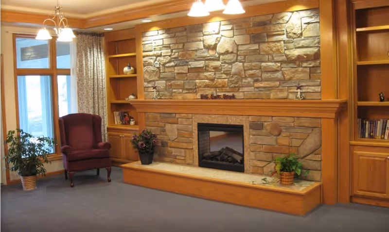 Cozy living room area with a stone fireplace surrounded by wooden built-in shelves and cabinets. There is a burgundy upholstered armchair near a window with curtains, and potted plants placed on the floor and the fireplace hearth. The room is softly lit by ceiling lights.