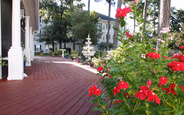 Red wooden deck patio with red flowering plants in the foreground, a white tiered fountain, and a multi-story building in the background.