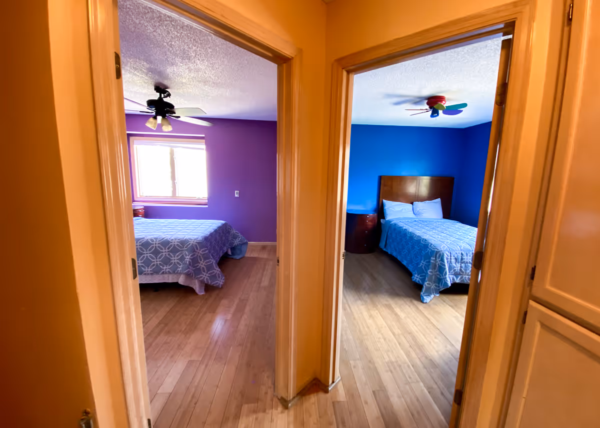 Hallway view into two adjacent bedrooms painted purple and blue, each with a bed, ceiling fan, and wood floors.
