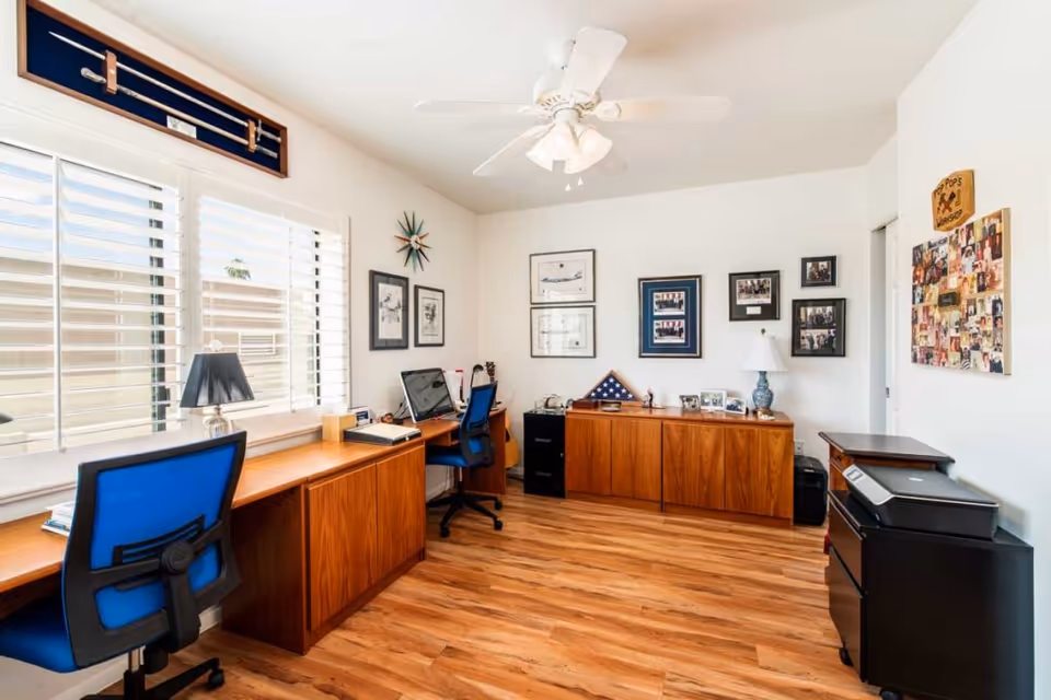 A bright office room with wooden flooring and white walls. The room features two blue office chairs, a long wooden desk under a window with white blinds, a computer, a lamp, and various framed pictures and decorations on the walls. There is also a wooden cabinet with more framed photos and a folded American flag, as well as a black printer on a small black cabinet.