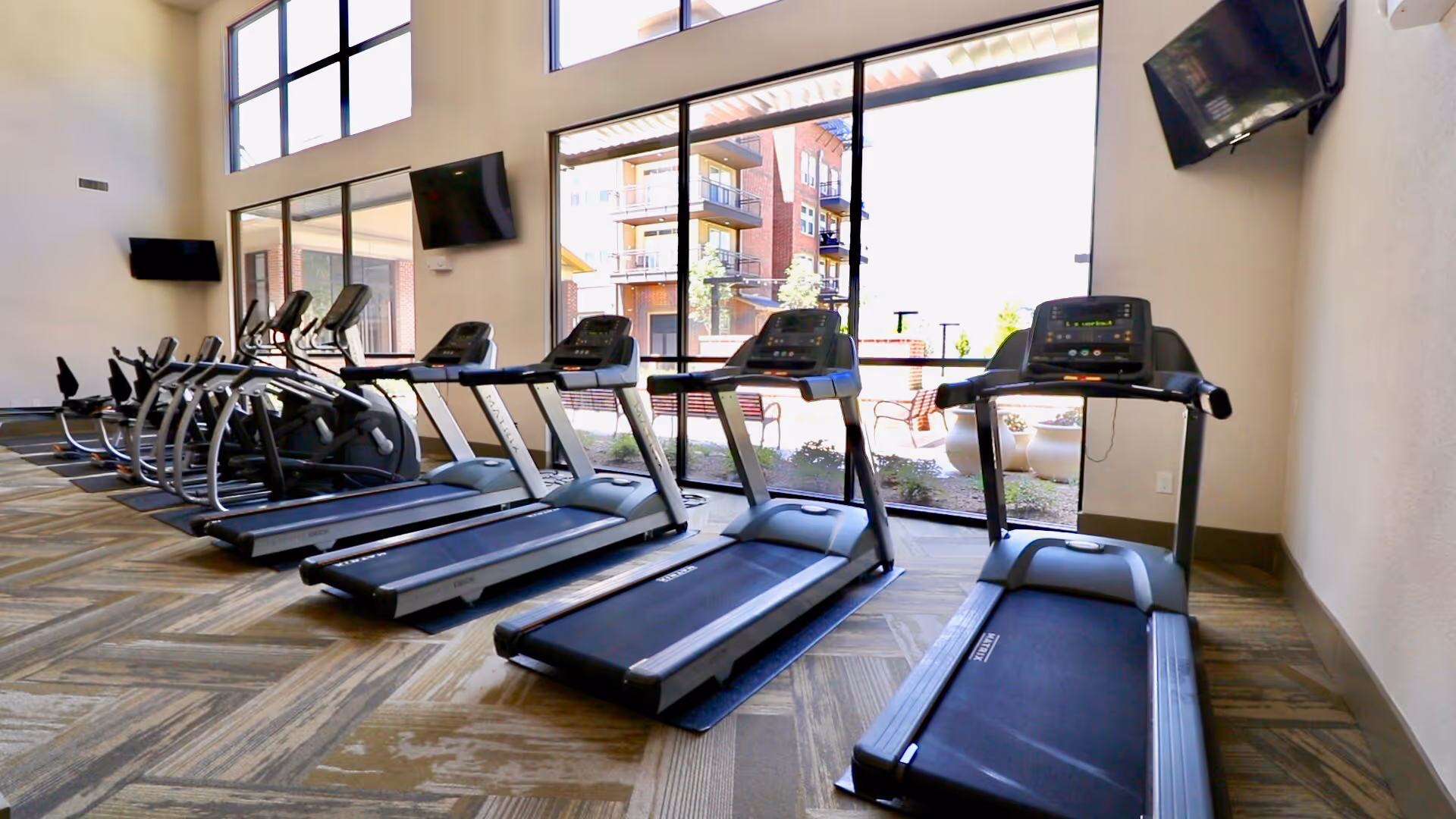 A fitness room with several treadmills and elliptical machines lined up facing large windows that let in natural light. Two wall-mounted televisions are visible, and outside the windows, there is a view of a patio area and neighboring buildings.