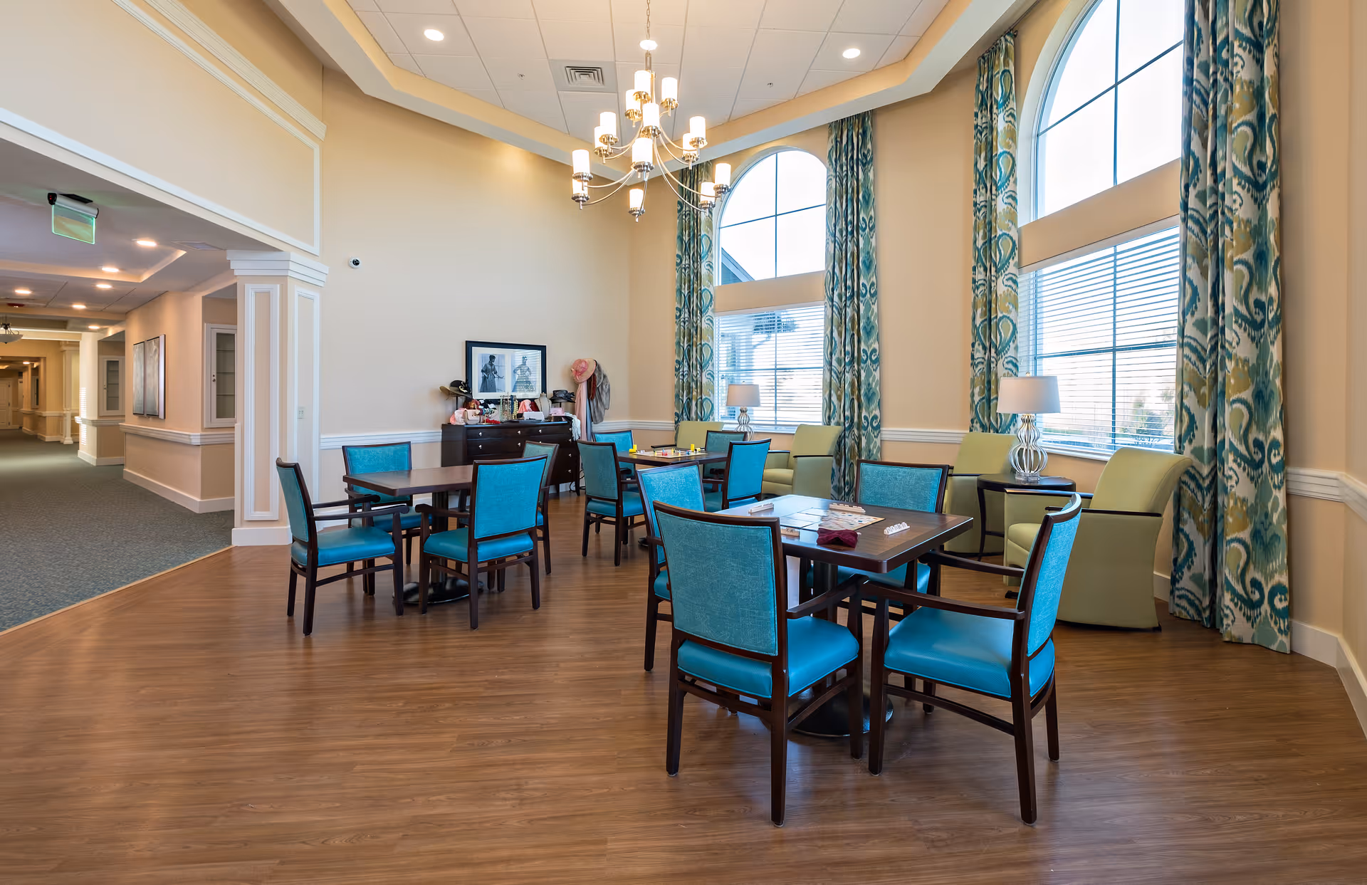 A bright and spacious common area in a senior living facility with several tables and turquoise chairs arranged for socializing or activities. Large arched windows with patterned curtains let in natural light. Comfortable green armchairs and side tables with lamps are placed near the windows. The room has wood flooring and a chandelier hanging from the ceiling. A hallway is visible to the left.