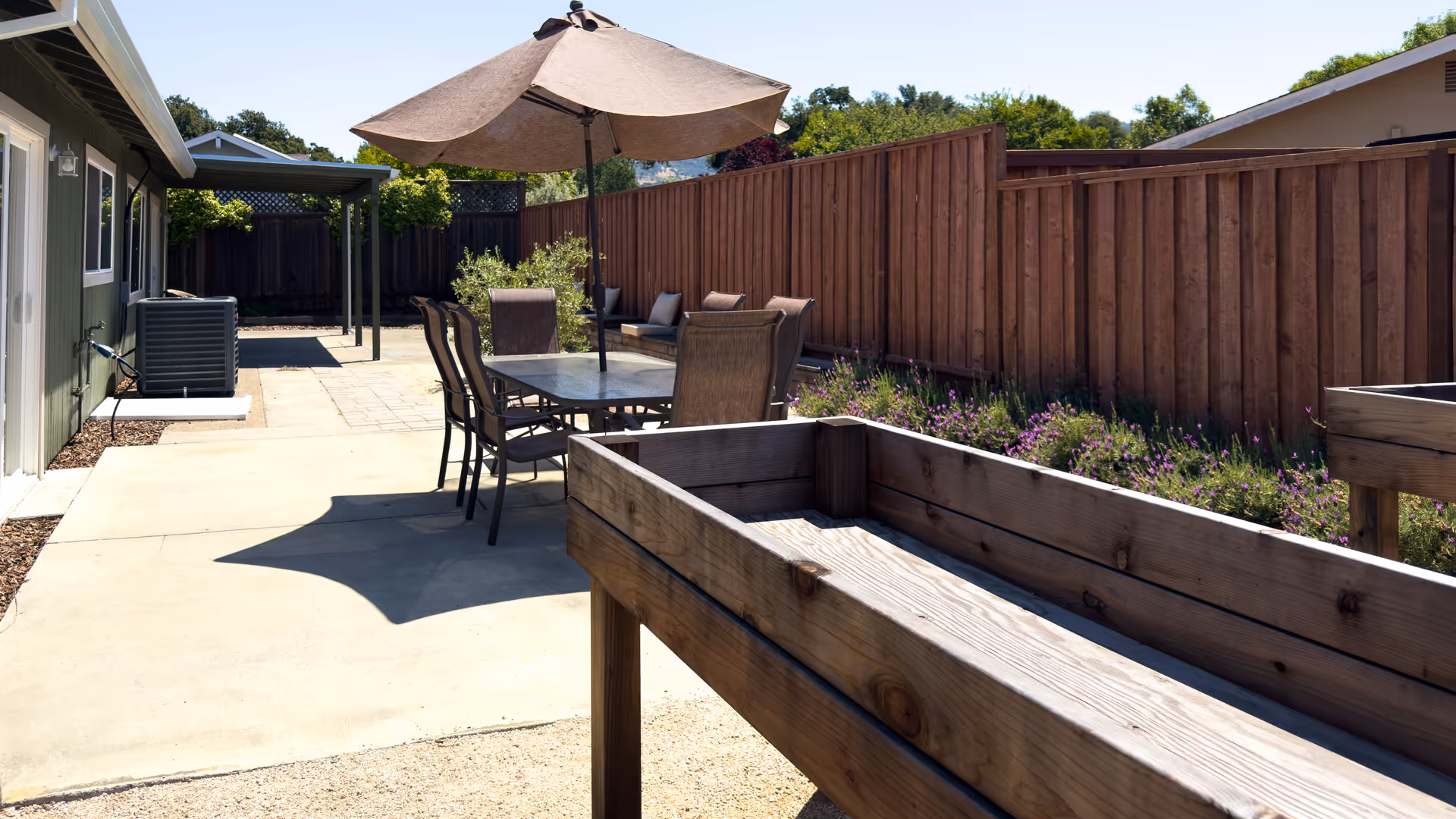 Sunlit outdoor patio with a table and umbrella, chairs, raised wooden planters, and a tall wooden fence.