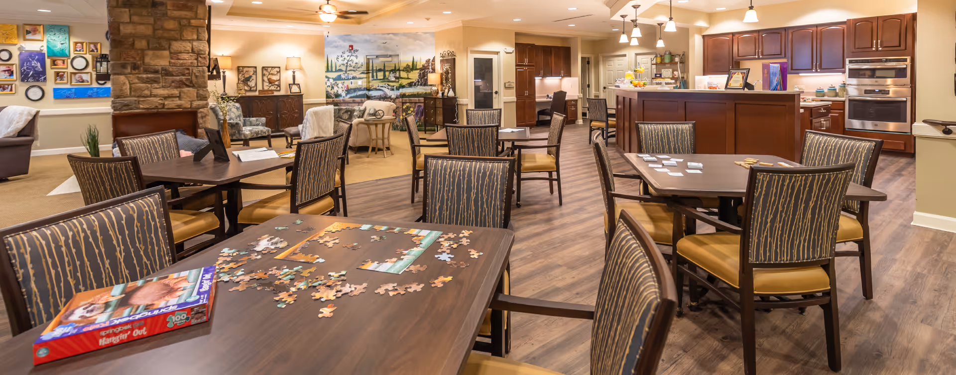 Spacious senior living common area with multiple dining tables and chairs, a kitchenette in the back, and a partially completed puzzle on the foreground table.