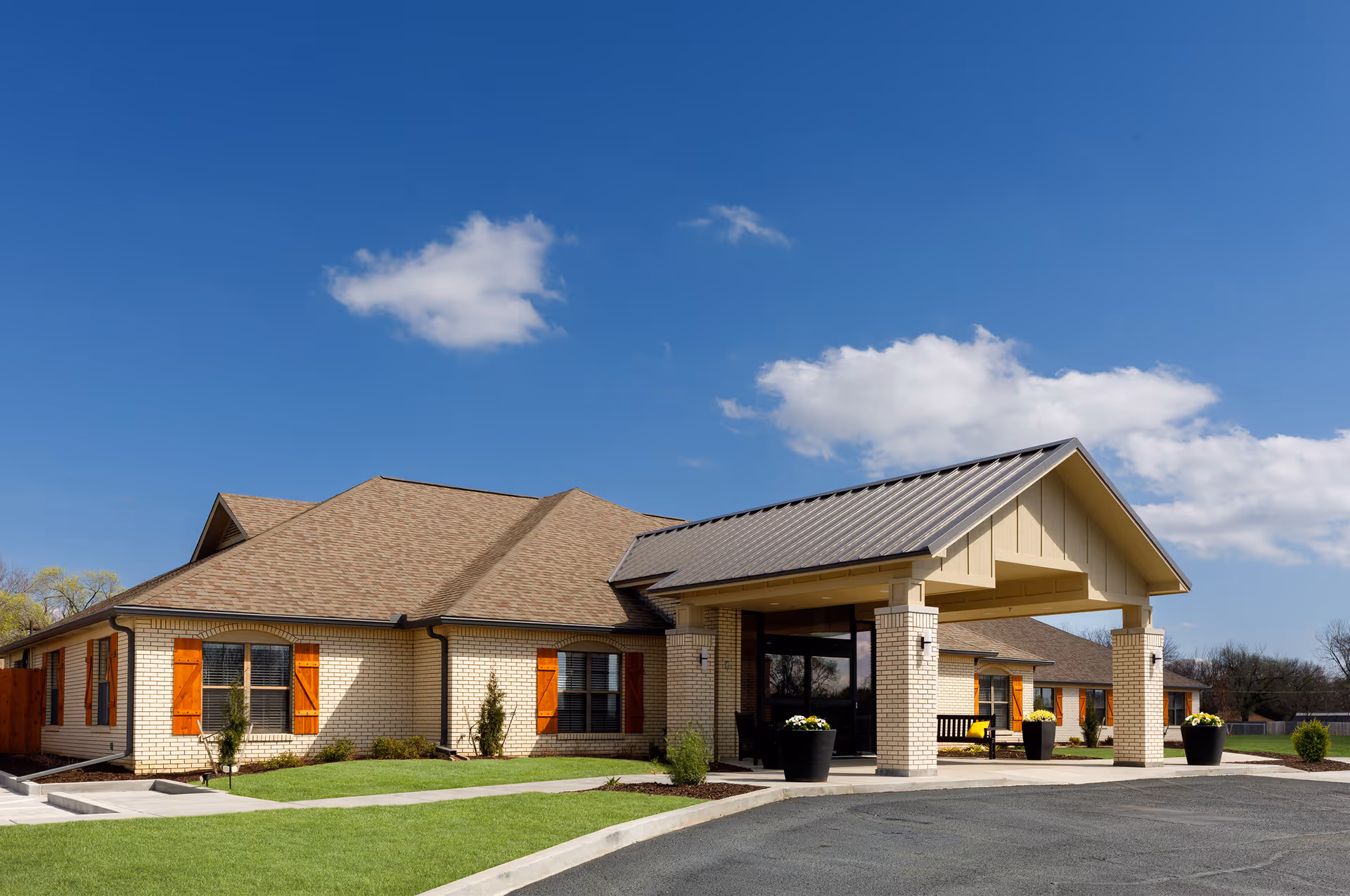 Exterior view of a single-story senior living facility building with beige brick walls, brown shingled roof, and a covered entrance supported by columns. The building has windows with wooden shutters, a well-maintained lawn, and a clear blue sky with a few clouds.
