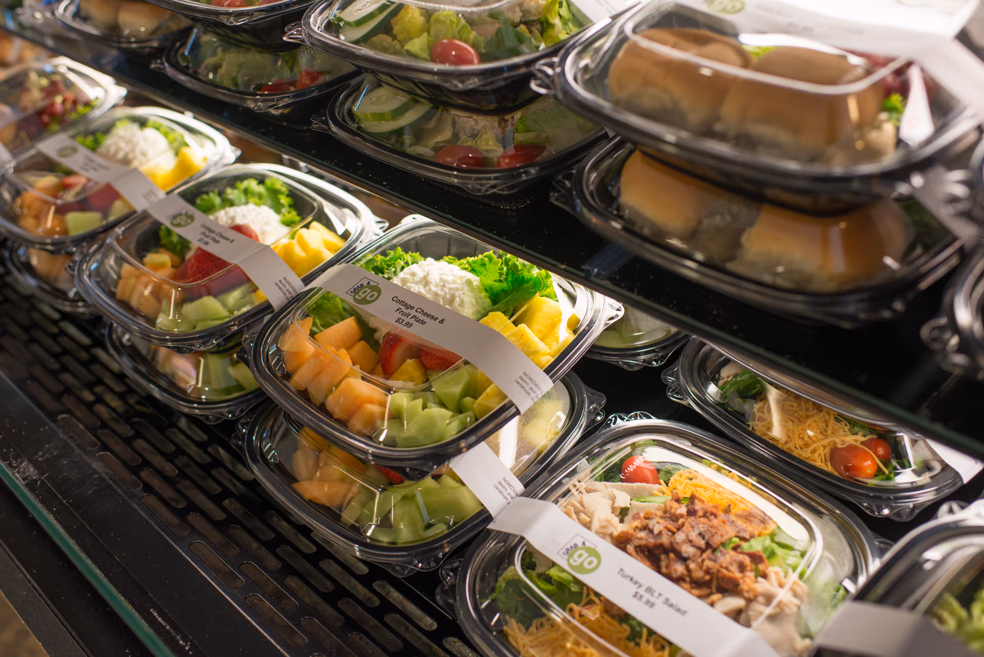 Pre-packaged salads and fruit bowls displayed in a refrigerated case, including cottage cheese and fruit plate and turkey BLT salad, ready for grab-and-go service.