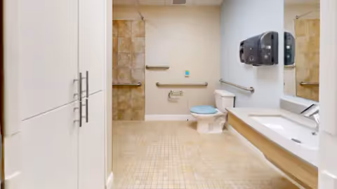 Accessible bathroom with beige tiled floor and walls, a white toilet with a blue seat, grab bars on the walls, a white sink with a large mirror above it, and a paper towel dispenser mounted on the wall.