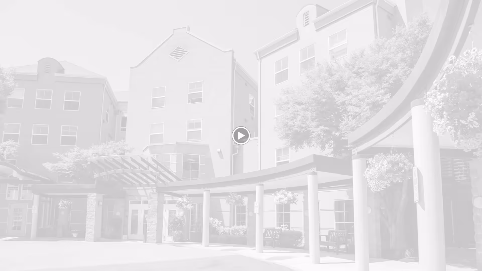Exterior front view of a multi-story retirement facility building with large windows, a covered entrance supported by columns, hanging flower baskets, and benches near the entrance.