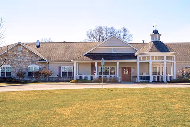 Exterior view of a single-story senior living facility building with a combination of stone and siding exterior, a covered entrance with white columns, and a small cupola on the roof. The building is surrounded by a well-maintained lawn and leafless trees in the background under a clear blue sky.