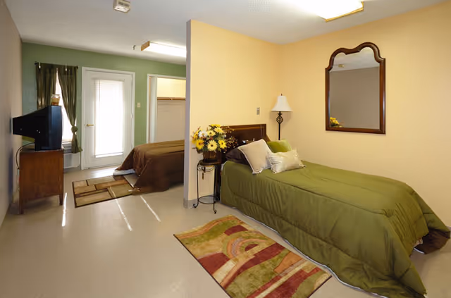 Interior view of a room in an assisted living facility showing two beds separated by a partial wall. The foreground bed has a green bedspread with pillows and a small side table with a flower arrangement and lamp. A mirror hangs on the wall above the bed. The background bed has a brown bedspread and is near a door with a window covered by curtains. A TV sits on a wooden stand to the left side of the room.