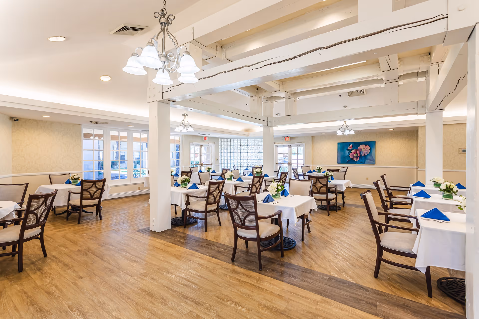 Bright and spacious dining room with multiple tables covered in white tablecloths, each set with blue folded napkins and small floral centerpieces. The room features wooden flooring, white beams on the ceiling, chandeliers, large windows, and a floral painting on the far wall.