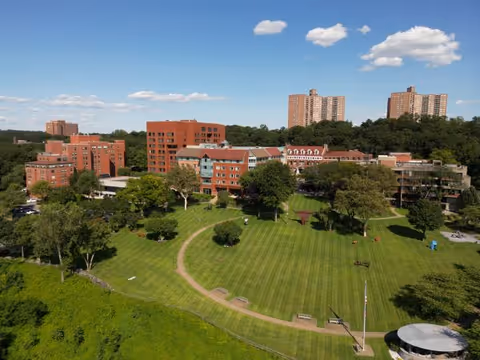 Aerial view of RiverSpring Assisted Living facility showing multiple red brick buildings surrounded by green lawns, trees, and walking paths under a blue sky with scattered clouds.