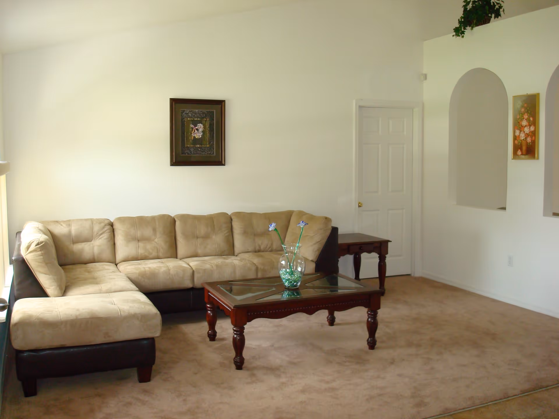A living room with a beige sectional sofa, a wooden coffee table with a glass top holding a vase with decorative flowers, beige carpet, white walls, a framed picture on the wall, and an arched wall opening with a floral painting.