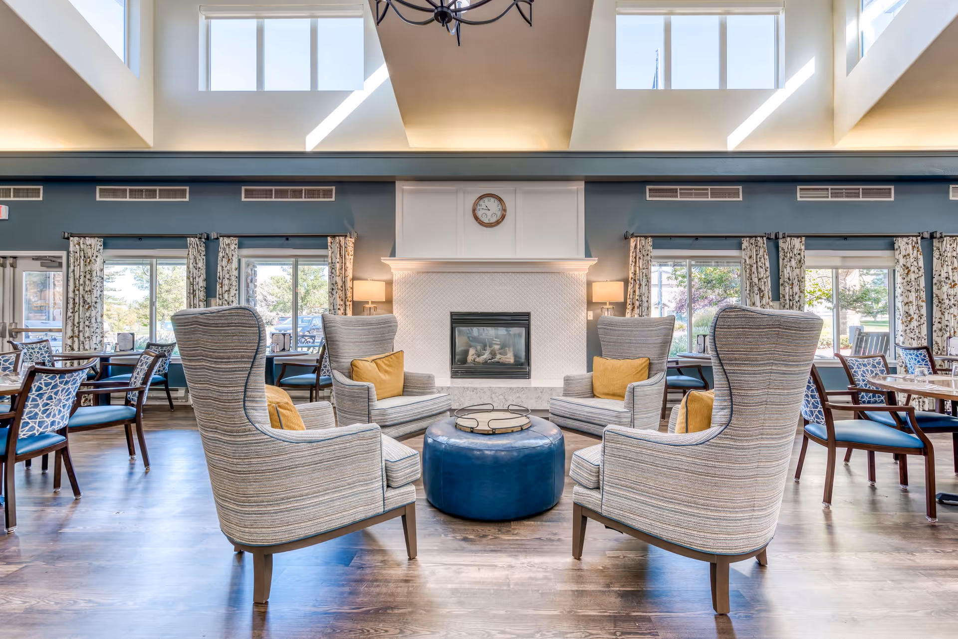 A bright and spacious senior living common area with four striped armchairs arranged around a round blue ottoman in front of a white fireplace. The room has large windows with patterned curtains, wooden floors, and several dining tables with chairs in the background. Natural light streams in from high windows near the ceiling.