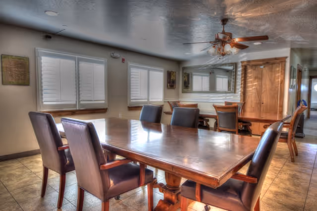 A well-lit dining room with a large wooden table surrounded by six dark cushioned chairs. The room has tiled floors, three windows with white shutters, a ceiling fan with lights, a large mirror on the wall, and wooden doors. The walls are decorated with framed artwork.