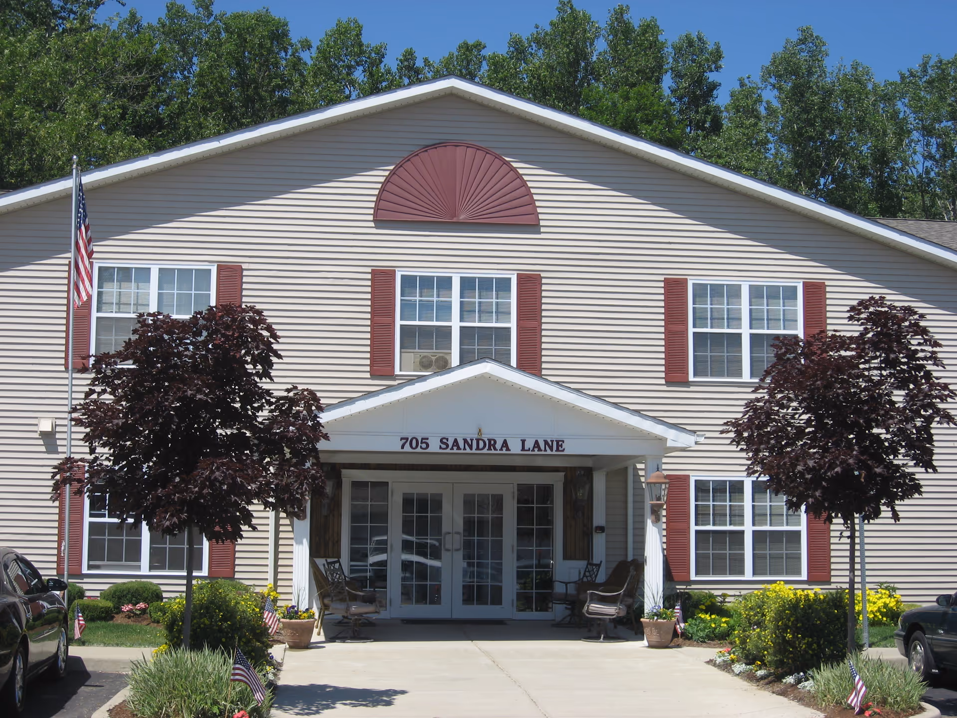 Front exterior view of a two-story beige building with red shutters and a white entrance canopy displaying the address 705 Sandra Lane. There are two small trees and landscaped bushes in front, with an American flag on a pole to the left and parked cars on either side of the driveway.