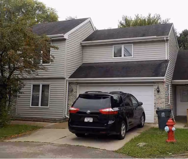 Exterior view of a two-story residential building with beige siding and a black roof. A black minivan is parked in the driveway in front of a white garage door. There is a red fire hydrant and a trash bin near the driveway, with some trees and grass around the property.