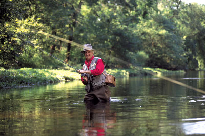 An elderly man wearing a hat and fishing vest stands waist-deep in a calm river, fly fishing with a fishing rod surrounded by lush green trees and vegetation.