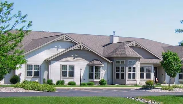 Exterior front view of a single-story assisted living building with gabled roofs, bay windows, lawn, and a driveway.