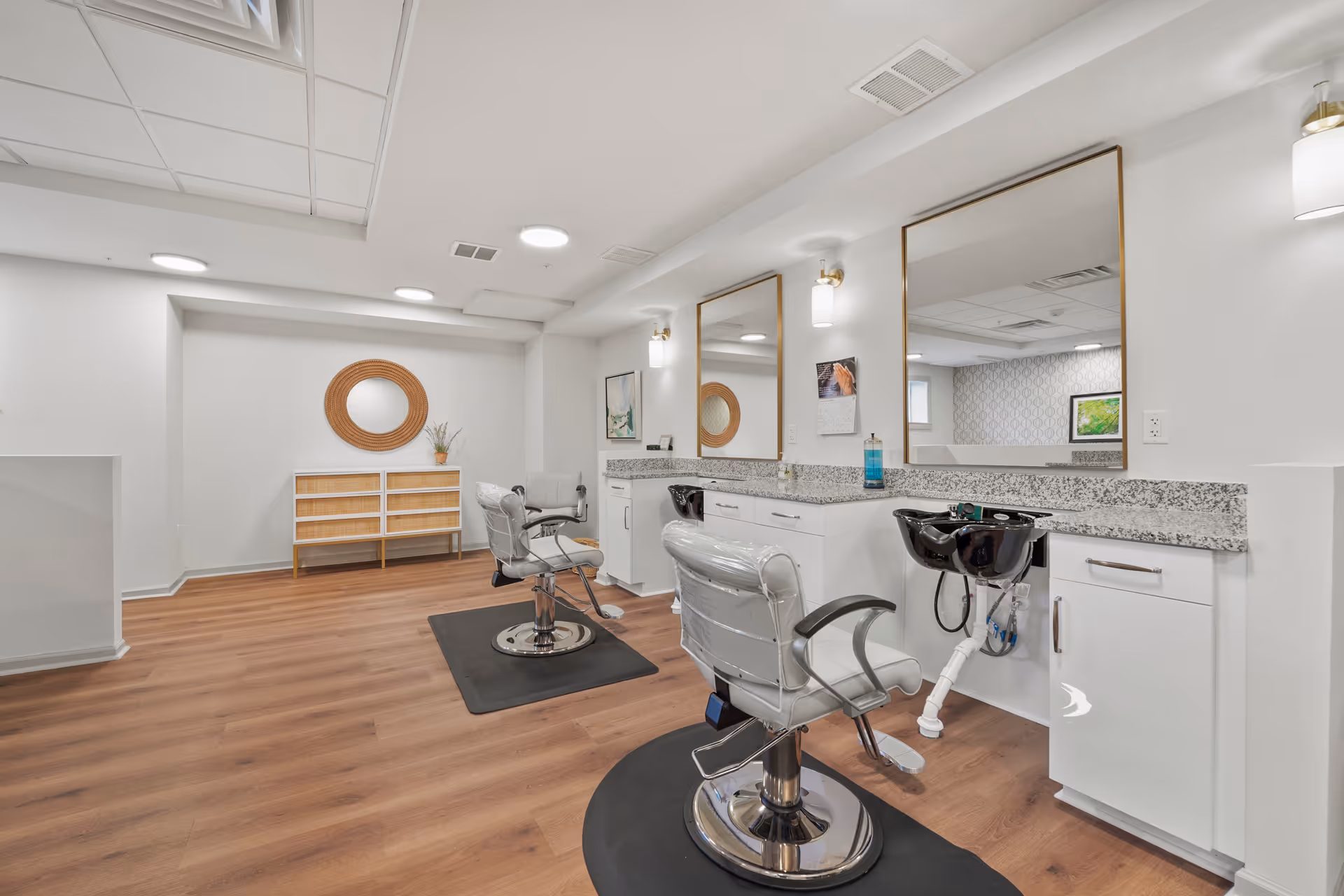 Interior view of a salon area in a senior living facility with two styling chairs in front of large mirrors, a granite countertop with a black wash basin, white cabinets, and wooden flooring. The room is well-lit with ceiling lights and wall sconces, and there is a decorative round mirror and a small cabinet against the far wall.
