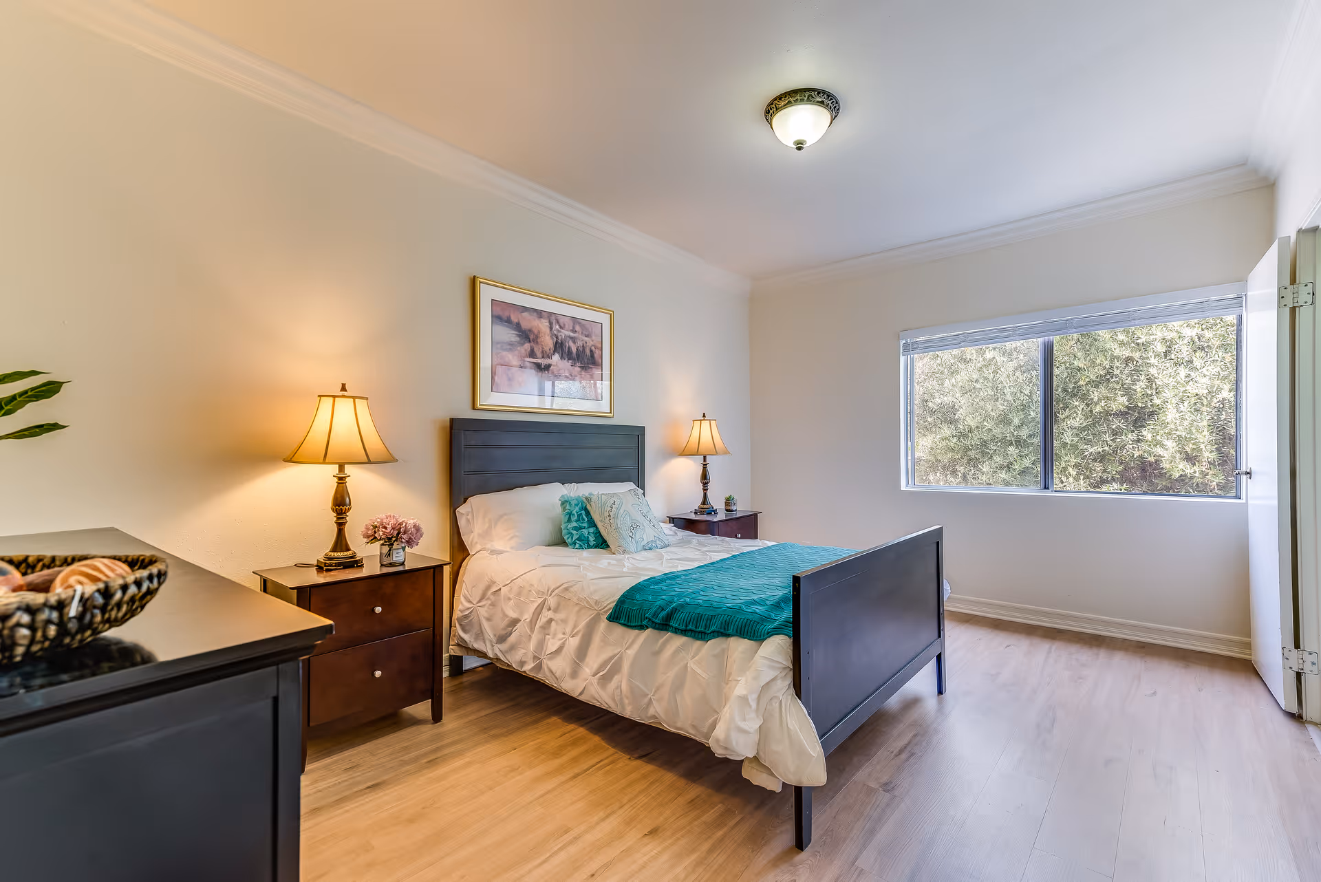 A bright and clean bedroom with a double bed featuring white bedding and teal accents. The room has wooden flooring, two bedside tables with lamps, a framed picture above the bed, a large window with a view of greenery outside, and a dresser with a decorative bowl.