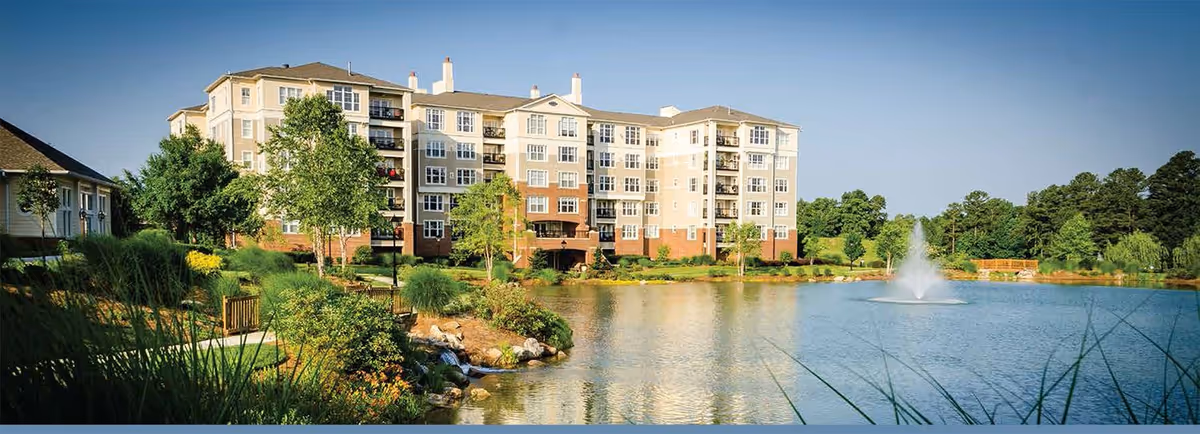 A large multi-story senior living facility building situated beside a pond with a water fountain. The area around the pond is landscaped with trees, shrubs, and grass under a clear blue sky.