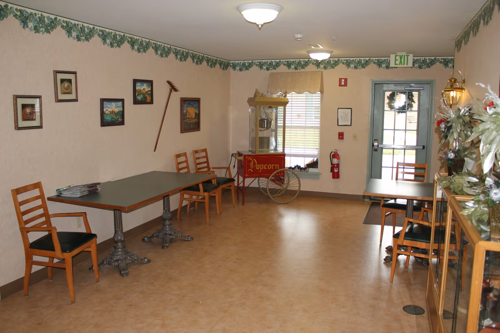 A small common area with two tables and wooden chairs along the walls. There is a vintage-style popcorn machine near a window with blinds. The walls are decorated with framed pictures and a leafy border near the ceiling. A door with a wreath and an exit sign is visible at the far end. There are some decorative plants on a glass display cabinet on the right side.