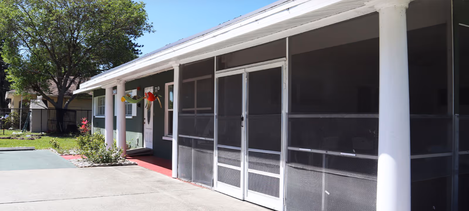 Single-story house with green siding, a screened front porch with white columns, and a driveway under a clear blue sky.