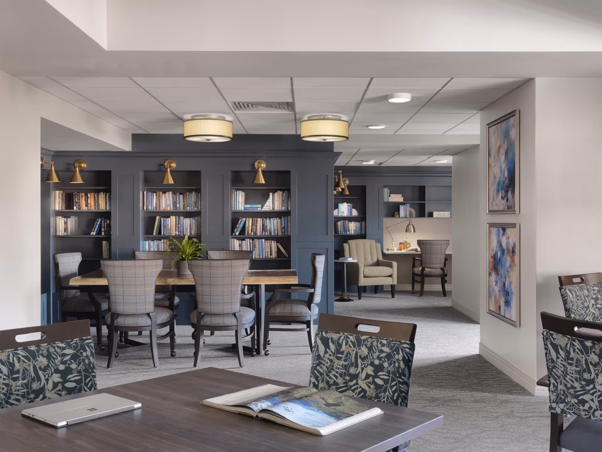 A well-lit interior common area with a large wooden table surrounded by six upholstered chairs in front of built-in dark blue bookshelves filled with books. There are brass wall lamps above the bookshelves. In the background, there is a cozy seating area with an armchair, a desk with a lamp, and another chair. The walls are decorated with two abstract paintings, and the floor is carpeted in a light gray tone. A table in the foreground has an open book and a closed laptop on it.