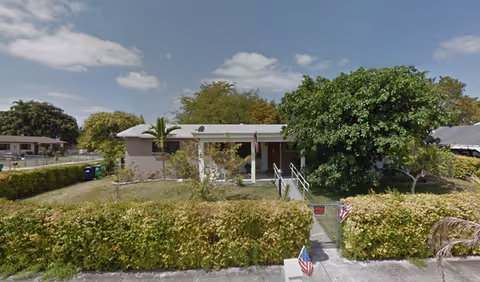 Single-story building with a light-colored exterior surrounded by green bushes and trees under a partly cloudy sky. There is a small ramp leading to the entrance, and American flags are displayed near the entrance and on the sidewalk. The area appears to be a residential or community setting.