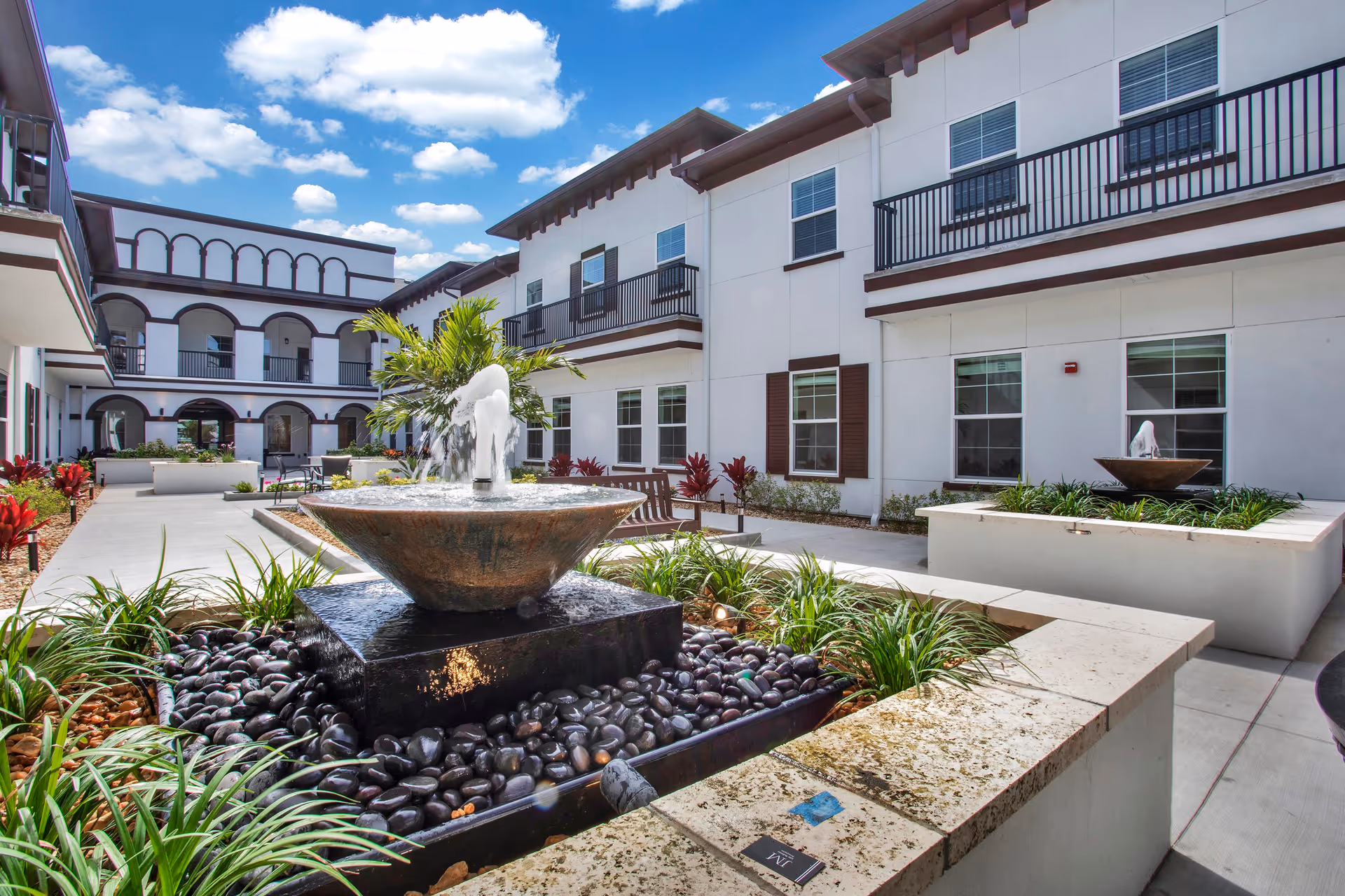 Outdoor courtyard area of The Blake at LPGA featuring two modern water fountains surrounded by plants and black decorative stones, with white buildings and balconies under a blue sky with scattered clouds.
