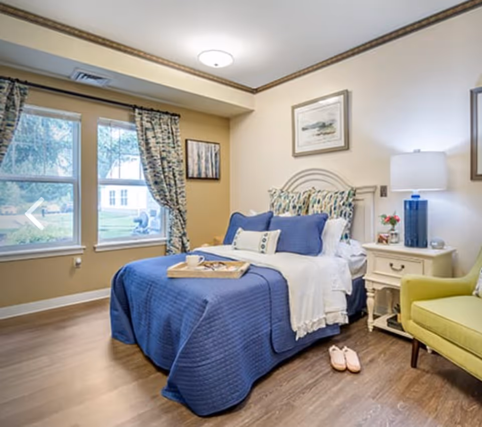 A cozy bedroom with a neatly made bed covered in blue bedding and multiple pillows. A wooden tray with a cup and glasses is placed on the bed. There are two windows with patterned curtains letting in natural light. A nightstand with a blue lamp, flowers, and other small items is beside the bed. A green armchair is positioned near the nightstand. The room has wooden flooring and light-colored walls with framed artwork.