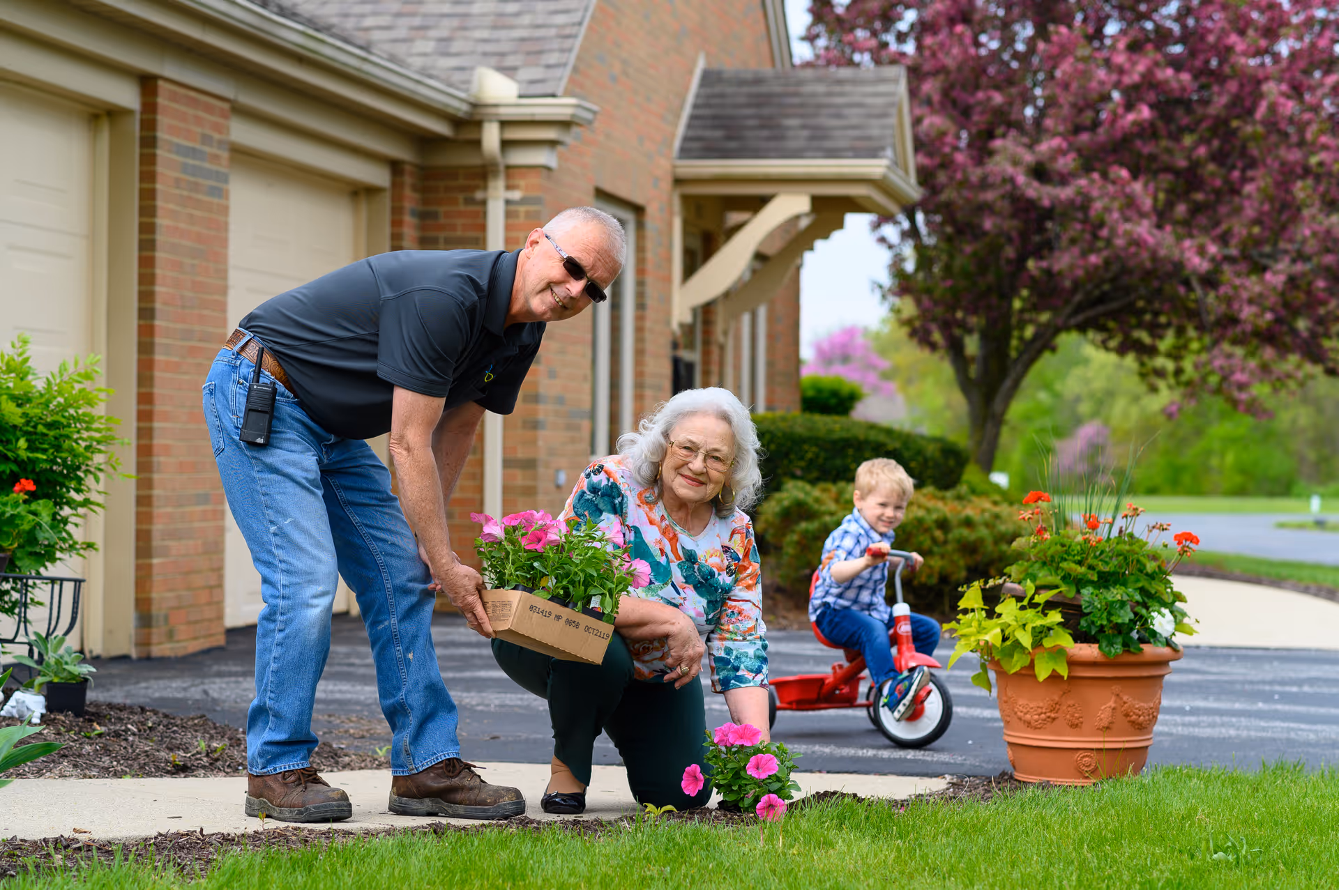 An elderly woman kneeling on the grass planting pink flowers while an elderly man holds a tray of pink flowers beside her. A young child rides a red tricycle on the driveway in the background. They are outside a brick building with a garage and blooming trees.