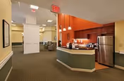 Interior view of a senior living facility showing a small kitchen area with a refrigerator, cabinets, and a counter with pendant lights above. The kitchen area opens into a hallway leading to a common seating area with chairs and tables. The walls are painted in warm tones with green lower panels and orange accent wall behind the kitchen.