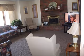 A cozy living room with a floral patterned sofa, two beige armchairs, a brick fireplace with decorative items on the mantel, a television on a stand showing a woman wearing sunglasses, a side table with a lamp, and framed pictures on the walls.