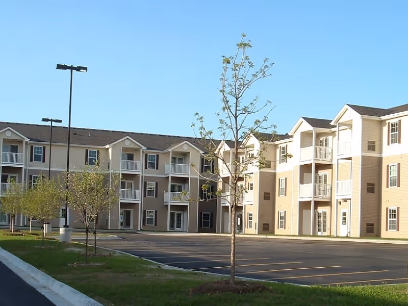 Exterior view of a three-story residential building with balconies, multiple windows, and a parking lot in front. Young trees and grass line the parking area under a clear blue sky.