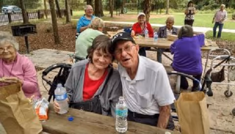 A group of elderly people sitting and standing around a wooden picnic table in an outdoor park setting. Some are smiling and appear to be enjoying each other's company. There are water bottles and brown paper bags on the table, and trees and a walking path are visible in the background.