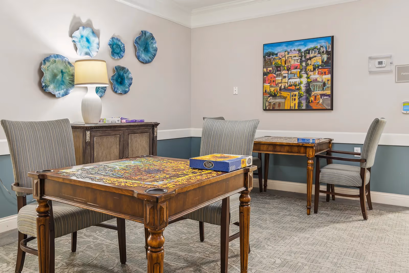 A cozy interior room with two wooden tables and four striped upholstered chairs. One table has a partially completed jigsaw puzzle and a puzzle box on it. The walls are decorated with blue ceramic wall art and a colorful painting of a cityscape. A white lamp sits on a wooden cabinet against the wall.