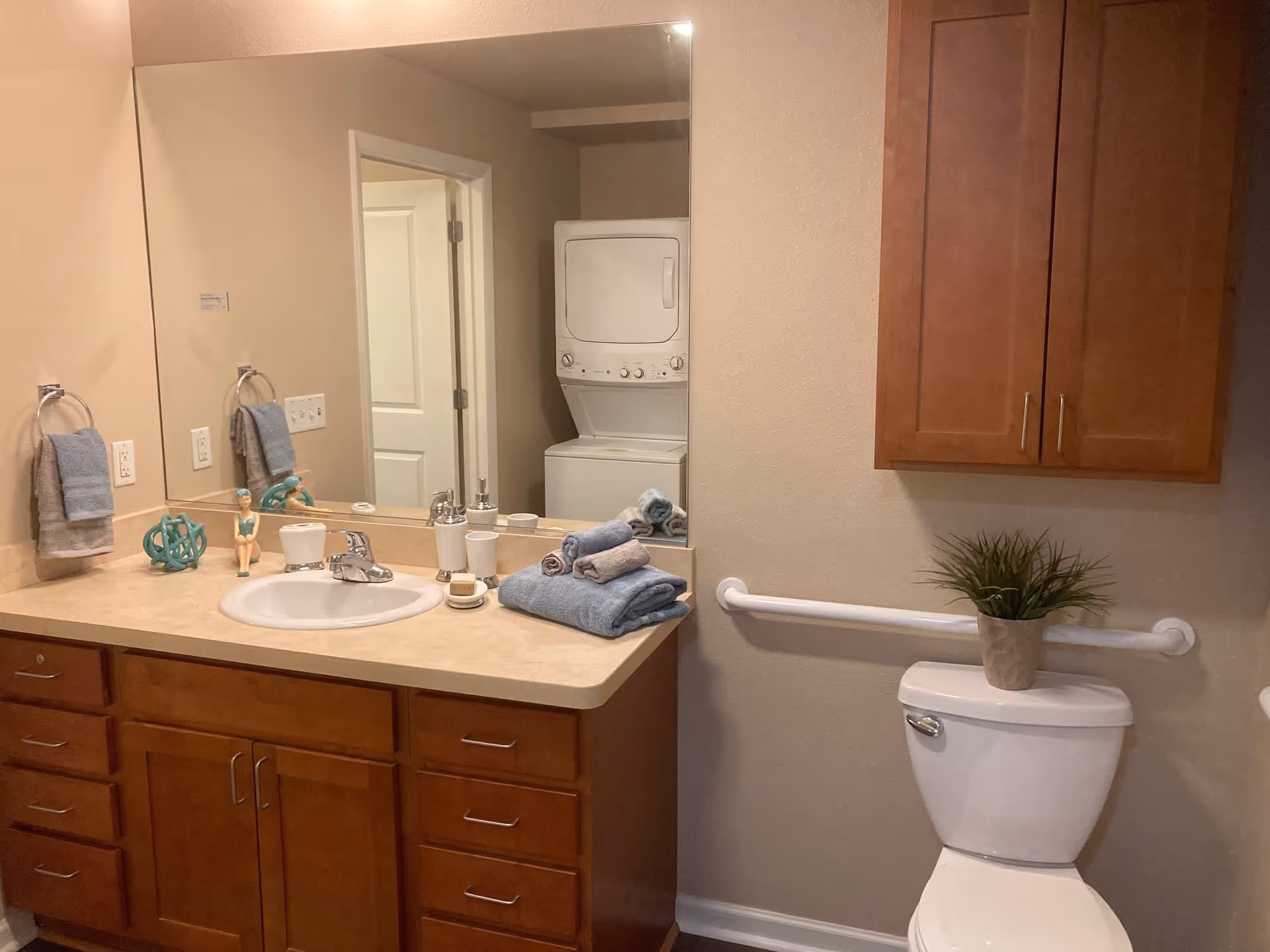 Bathroom with a large mirror above a countertop sink, wooden cabinets below, and a toilet with a potted plant on top. A towel ring with a blue towel is mounted on the wall next to the sink. A stackable washer and dryer unit is visible through an open doorway reflected in the mirror.