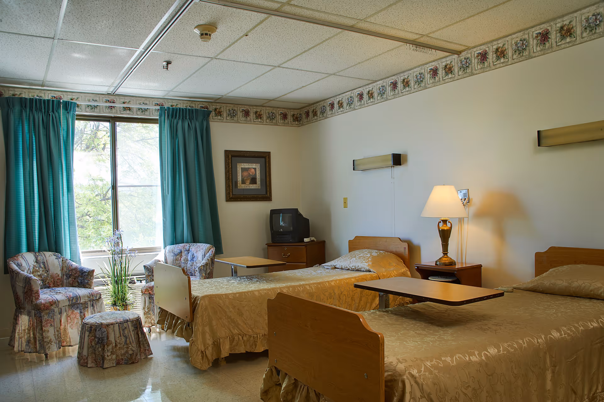 A senior living facility bedroom with two single beds covered in gold bedspreads. Each bed has an overbed table. There is a nightstand with a lamp between the beds. Two floral upholstered armchairs and a matching ottoman are placed near a window with teal curtains. A small TV sits on a wooden dresser, and a framed picture hangs on the wall. The room has a floral border near the ceiling.