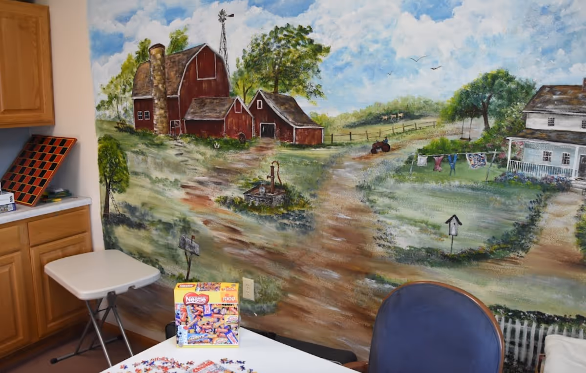 A room corner with a painted mural of a rural farm scene featuring red barns, a windmill, a dirt path, trees, a tractor, and a farmhouse with a porch and clothesline. In the foreground, there is a white folding table with a Nestle puzzle box and puzzle pieces, a blue chair, a small white folding table, and wooden cabinets on the left side.