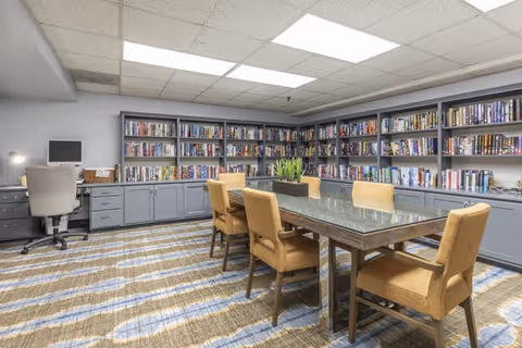 A well-lit library or reading room with gray bookshelves filled with books lining the walls. In the center, there is a rectangular table with a glass top surrounded by six mustard-yellow upholstered chairs. A small green plant is placed on the table. To the left, there is a desk with a computer and an office chair. The room has a patterned carpet and a drop ceiling with fluorescent lights.