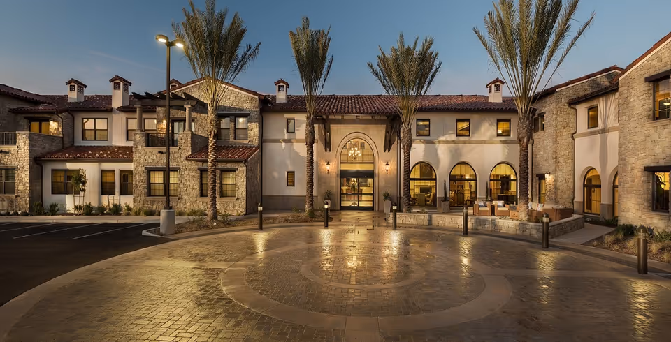 Front exterior of a Mediterranean-style two-story senior living building with arched entrance, palm trees and a circular driveway at dusk.
