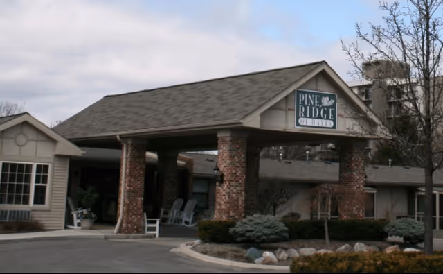 Entrance of a senior living facility with a covered driveway supported by brick pillars. The sign on the building reads 'Pine Ridge of Davis'. There are some shrubs, trees, and a curved driveway in front of the entrance.