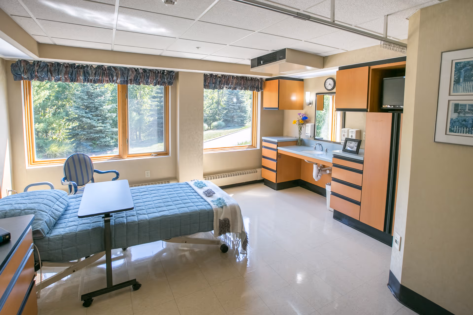 A bright and clean patient room with a hospital bed covered in a blue quilt, a small overbed table, a striped armchair, large windows with a view of green trees outside, and a built-in wooden cabinet with a sink, mirror, and small TV.