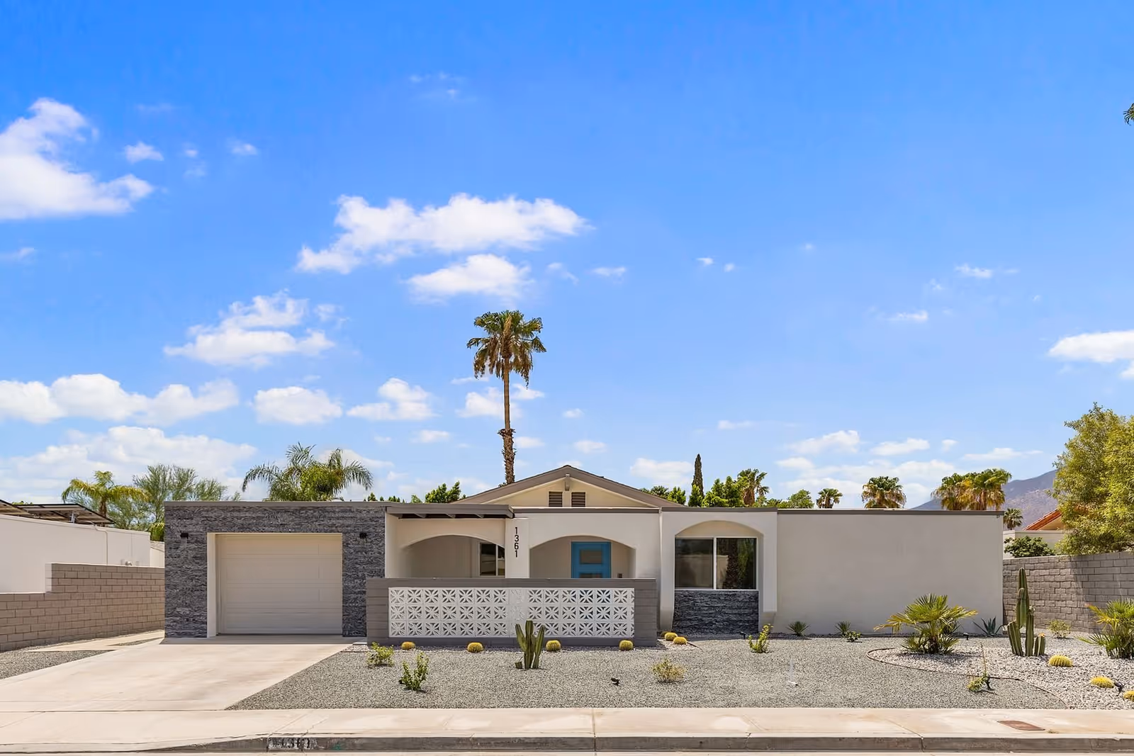 Single-story modern stucco building with a garage, decorative breeze block wall, desert landscaping and a tall palm tree under a bright blue sky.
