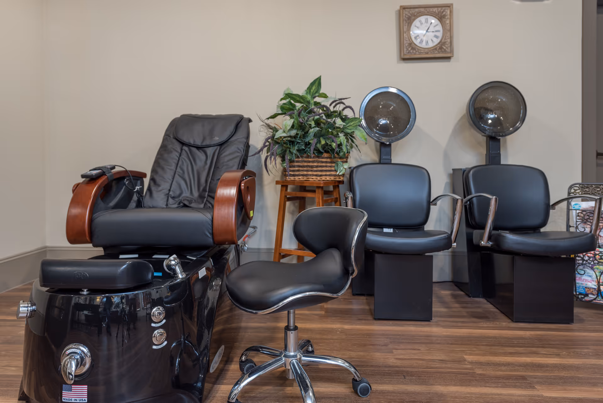 Salon area with a pedicure chair, rolling stylist stool, two hooded hair dryers, and a potted plant against a neutral wall.