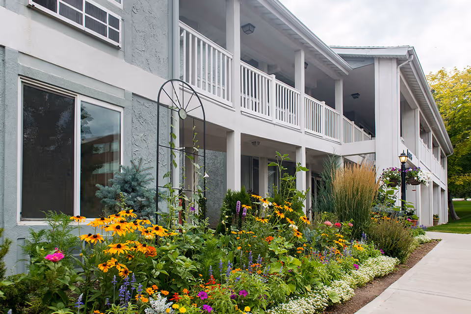 Exterior view of a two-story building with white railings and a garden full of colorful flowers and greenery along the walkway at Highland Cove Retirement.