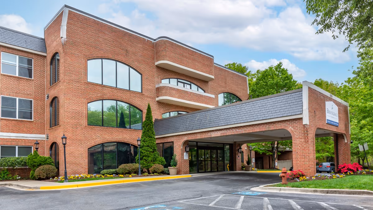 Exterior view of a three-story brick building with large arched windows and a covered entrance driveway. The building is surrounded by landscaped greenery, including bushes, trees, and colorful flowers. A sign on the covered entrance reads 'Brookdale Potomac.'